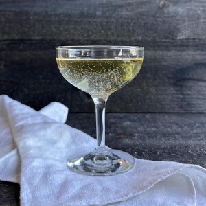May include: A coupe glass filled with a sparkling, golden-colored beverage. The glass sits on a white linen napkin against a dark wood background. Bubbles rise in the drink, suggesting effervescence.