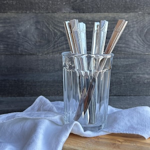 May include: A clear glass with silver silverware inside, resting on a wooden cutting board and white cloth. The silverware has decorative patterns. The background is a dark wood surface.