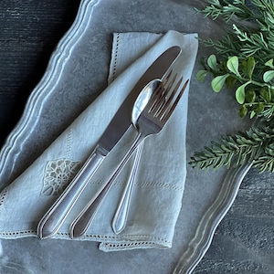 May include: A silver-toned metal tray with a scalloped edge holds a cream-colored linen napkin featuring delicate lace trim and embroidery. A stainless steel dinner knife, spoon, and fork are arranged neatly on top of the folded napkin.