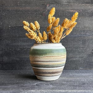 May include: A ceramic vase with a striped design in shades of green, brown, and beige, holding dried yellow flowers. The vase has a rounded shape and a white base. The background is a dark wood surface.