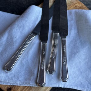 May include: A collection of silver-colored knives with dark blades, arranged on a white cloth and a wooden cutting board. The handles feature a decorative floral design. The knives are likely for dining and add a touch of elegance to any table setting.