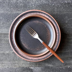 May include: A dark brown plate with a reddish-brown rim and a silver fork with a wooden handle. The plate is centered on a dark gray wooden surface.