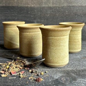 May include: Four cylindrical ceramic cups with a speckled mustard yellow glaze and flared rims. The cups have a smooth, matte finish and are arranged in a group. A silver-toned metal spoon rests near a small pile of loose-leaf herbal tea.