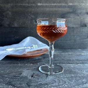 May include: A clear glass coupe cocktail glass featuring a delicate etched leaf pattern around the bowl, filled with an amber-colored liquid. The stem is faceted and leads to a circular base, showcasing a refined design for serving beverages.