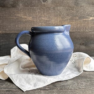 May include: A blue ceramic pitcher with a handle, set on a white cloth. The pitcher has a rounded body and a spout. The background is a dark wooden surface.