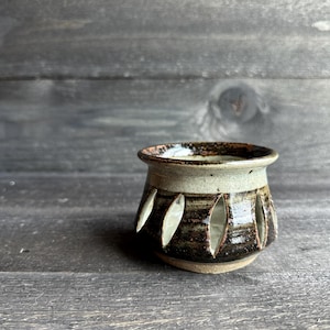 May include: A handmade ceramic cup with a unique design. The cup features a light tan rim and a dark brown body with leaf-shaped cutouts. The interior is dark brown, and the base is a lighter tan color. The cup sits on a wooden surface.