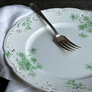 May include: A white ceramic plate with a scalloped edge and green floral patterns, featuring embossed scrollwork details along the rim. A silver-toned metal fork with an ornate handle and a decorative engraving on the base rests on the plate.