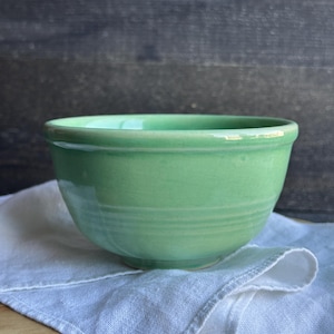 May include: A light green ceramic bowl with a rounded shape. The bowl sits on a white linen napkin. The background is a dark wooden surface. The bowl has a smooth, glossy finish.