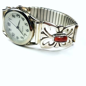 Op de afbeelding: Een zilveren horloge met een witte wijzerplaat en zwarte cijfers. De horlogeband is een zilveren uitbreidbare band met een vlinderontwerp en een rode steenaccent.