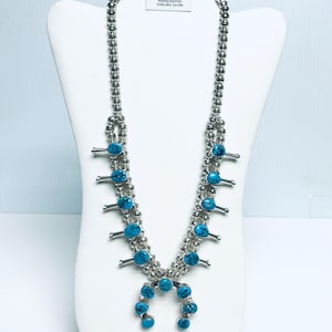 Könnte beinhalten: Eine silberne Squash Blossom Halskette und passende Ohrstecker mit Türkissteinen. Die Halskette wird auf einer weißen Büste präsentiert. Die Ohrringe und die Halskette sind mit "GENUINE INDIAN HANDCRAFTED STERLING SILVER" beschriftet.