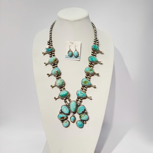 Puede incluir: Un collar squash blossom y pendientes a juego con piedras de turquesa pulidas engastadas en plata. El collar consta de cuentas de plata redondas y múltiples flores con turquesas que conducen a un colgante naja central.