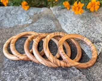 May include: A set of five circular wooden bangles featuring a natural light brown grain pattern with darker brown streaks. The bracelets are arranged in a slightly overlapping row, showcasing the smooth, polished finish of the wood material.