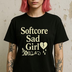 Pode incluir: Camiseta preta com o texto "Softcore Sad Girl" em letras brancas. Abaixo do texto, há um coração partido com sangue pingando. Abaixo do coração, há um desenho floral.