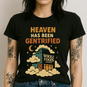 May include: Black t-shirt with the text "HEAVEN HAS BEEN GENTRIFIED" in orange and the Whole Foods Market logo. The design includes a building, clouds, a crescent moon, and stars.