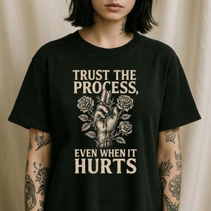 Op de afbeelding: Zwart t-shirt met de tekst "TRUST THE PROCESS, EVEN WHEN IT HURTS". Het ontwerp toont een hand met doornen en rozen. Het shirt heeft een ronde hals en korte mouwen.