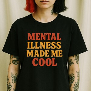 May include: Black t-shirt with the text "MENTAL ILLNESS MADE ME COOL" in orange and red block letters. The shirt has a crew neck and short sleeves. The overall theme is mental health awareness.
