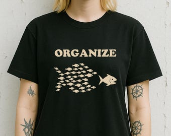 May include: Black t-shirt with the word "ORGANIZE" in a tan font. Below the text is a graphic of a school of small fish swimming towards a larger fish. The shirt is a crew neck style.