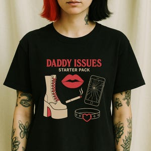 May include: Black t-shirt with the text "Daddy Issues Starter Pack" in red and white. The design includes a red lip, a broken phone, a cigarette, a platform boot, and a heart-shaped choker.