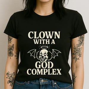 May include: Black t-shirt with white text that reads "Clown with a God Complex" and a black and white illustration of a clown with devil wings and a halo.