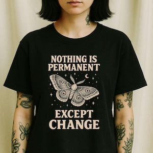 Op de afbeelding: Zwart t-shirt met de tekst "NOTHING IS PERMANENT EXCEPT CHANGE" en een illustratie van een mot. De mot is gedetailleerd met oogachtige markeringen en omgeven door sterren en een halve maan. De tekst en de mot zijn lichtbeige.