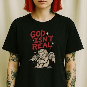 May include: Black t-shirt with the text "GOD ISN'T REAL" in red, dripping font. Below the text is a distressed illustration of a crying angel with wings. The shirt is a crew neck style.