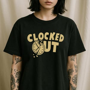 May include: Black t-shirt featuring the text "CLOCKED OUT" in gold. The letter "O" in "CLOCKED" is replaced with a broken clock graphic. The shirt has short sleeves.