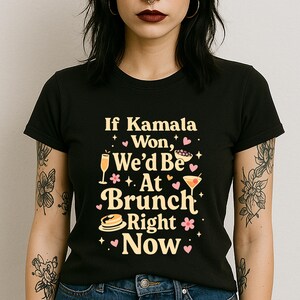 May include: Black t-shirt with the text "If Kamala Won, We'd Be At Brunch Right Now" in a playful font. The design includes illustrations of a champagne flute, a cocktail, pancakes, and hearts.