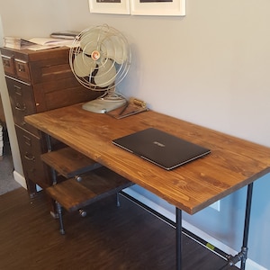 May include: A wooden desk with a black metal frame. The desk has two shelves and a laptop on the top surface. A fan is on the left side of the desk. The desk is in front of a wall with a white trim and a white thermostat. The wall also has four framed prints with black and white drawings of animals.