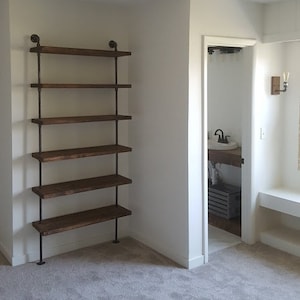 May include: A white room with a wooden shelf unit with five shelves. The shelf unit is made of metal pipes and wooden planks. The shelf unit is in the corner of the room. The room has white walls and a gray carpet.