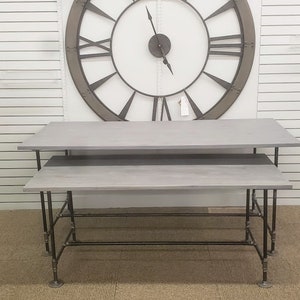 May include: Two gray wooden tables with black metal pipe legs. The tables are stacked on top of each other. A large, round, metal clock with Roman numerals is on the wall behind the tables.