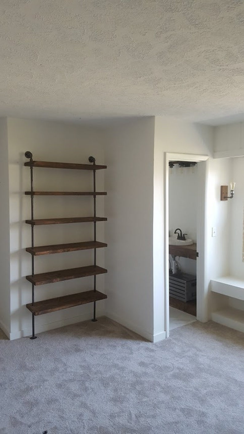 May include: A white room with a wooden shelf unit with five shelves. The shelf unit is made of metal pipes and wooden planks. The shelf unit is in the corner of the room. The room has white walls and a gray carpet.