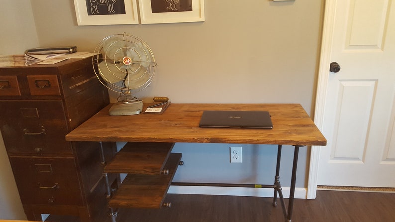 May include: A wooden desk with a metal pipe frame and three shelves. A laptop is on the desk and a metal fan is on the left side of the desk. The desk is in front of a white door and a gray wall.