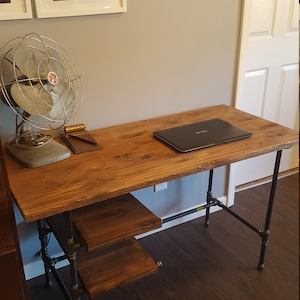 May include: A wooden desk with a black metal frame. The desk has a laptop on the top and a small shelf underneath. The desk is in a room with a light brown wood floor.