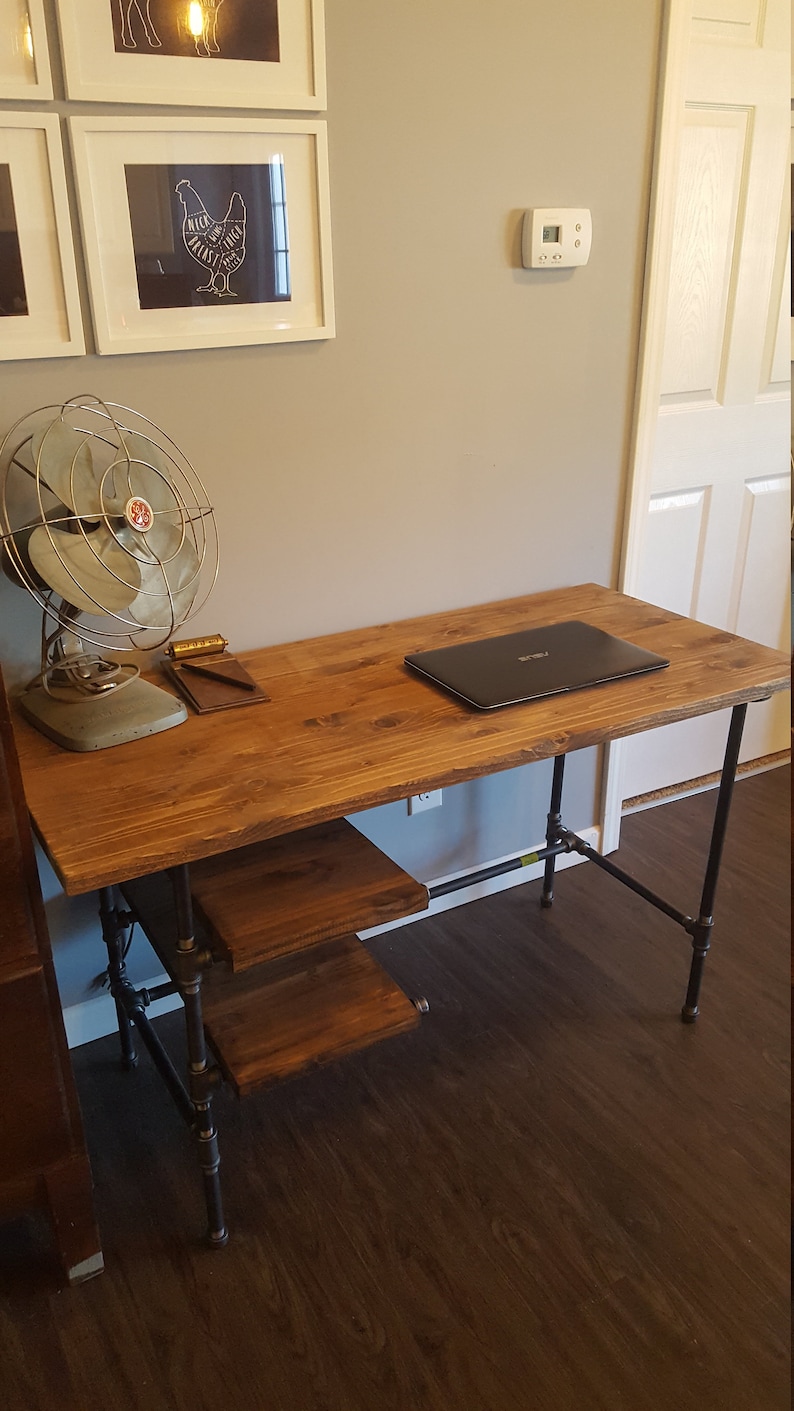 May include: A wooden desk with a black metal frame. The desk has a laptop on the top and a small shelf underneath. The desk is in a room with a light brown wood floor.