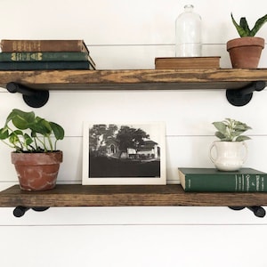 May include: Two wooden shelves with black metal brackets, holding books, plants, and a framed photo. The shelves are mounted on a white wall.