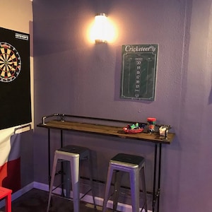 May include: A wooden bar table with black metal pipe legs and two silver metal bar stools. The table has a wooden top and a black metal pipe frame. The table is set against a purple wall with a dartboard and a chalkboard with the word "Cricketeer" on it.