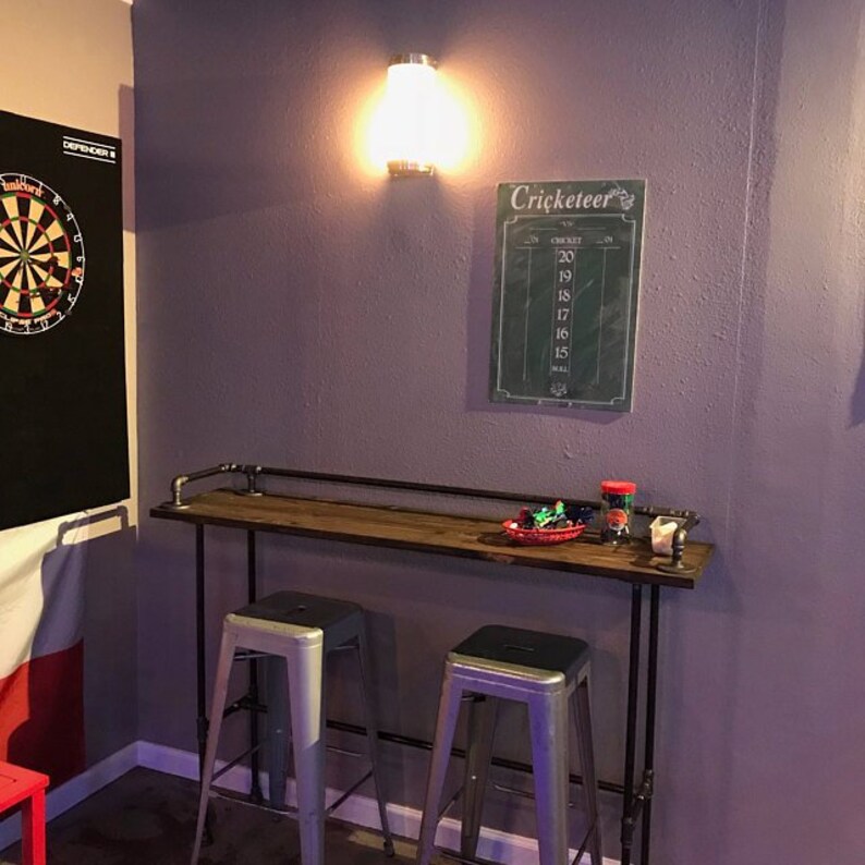 May include: A wooden bar table with black metal pipe legs and two silver metal bar stools. The table has a wooden top and a black metal pipe frame. The table is set against a purple wall with a dartboard and a chalkboard with the word "Cricketeer" on it.