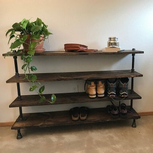 May include: A four-tier industrial pipe shelf unit with wooden shelves. The shelves are stained dark brown and the pipes are black. The shelf is holding a variety of items, including shoes, a plant, and a bag.