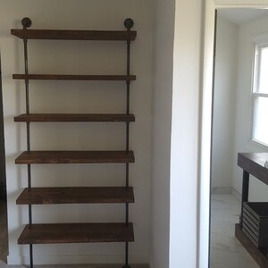May include: A brown wooden shelf unit with five shelves supported by black metal pipes. The shelf unit is in a room with white walls and carpeted floors.