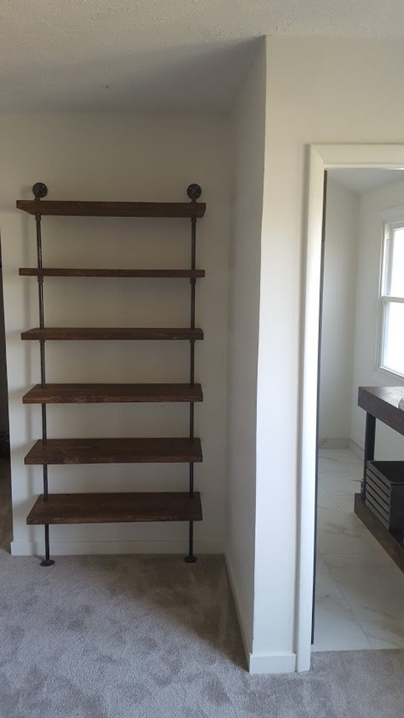 May include: A brown wooden shelf unit with five shelves supported by black metal pipes. The shelf unit is in a room with white walls and carpeted floors.