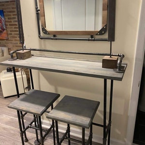 May include: A gray wooden bar table with black metal pipe legs and two matching gray wooden stools with black metal pipe legs. The bar table has a mirror above it with a wooden frame.