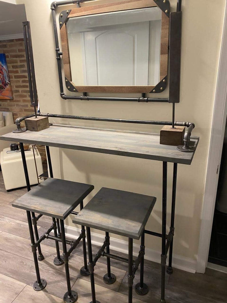 May include: A gray wooden bar table with black metal pipe legs and two matching gray wooden stools with black metal pipe legs. The bar table has a mirror above it with a wooden frame.