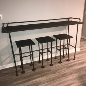 May include: A black metal bar table with a wooden top and three black metal stools. The table and stools are made of industrial pipe.