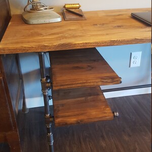 May include: A wooden desk with a metal pipe leg and a shelf underneath. The desk has a light brown wood top and a dark brown wood shelf. The desk is in front of a white wall with a light brown wood floor.