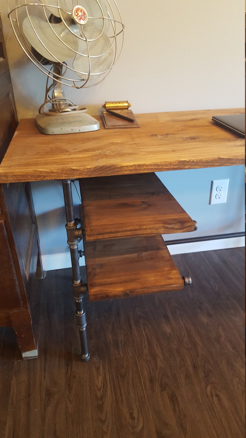 May include: A wooden desk with a metal pipe leg and a shelf underneath. The desk has a light brown wood top and a dark brown wood shelf. The desk is in front of a white wall with a light brown wood floor.