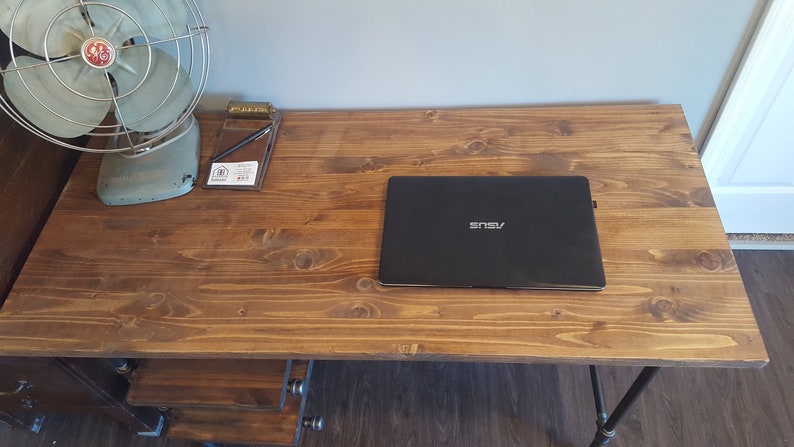 May include: A wooden desk with a black laptop computer on top of it. The desk has a brown wood top and black metal legs. There is a vintage fan and a notepad with a pen on the desk.