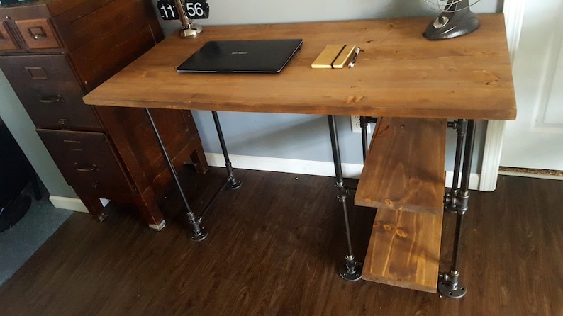 K&ouml;nnte beinhalten: Ein Holzschreibtisch mit schwarzen Metallrohrbeinen und einem unteren Regal. Der Schreibtisch hat einen Laptop und ein Notizbuch auf der Oberfl&auml;che.