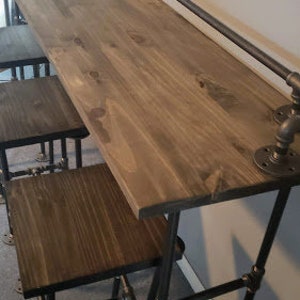 May include: A wooden countertop with a black metal pipe frame and two matching bar stools. The countertop is made of wood planks and has a rustic finish. The stools are made of black metal pipe and have a simple design.