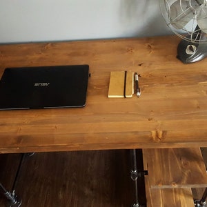 K&ouml;nnte beinhalten: Ein Holztisch mit schwarzen Metallbeinen. Auf dem Tisch liegen ein Laptop, ein Notizbuch und ein Stift. Links auf dem Tisch steht eine Uhr, die 11:56 Uhr anzeigt.