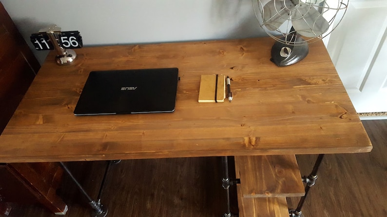 K&ouml;nnte beinhalten: Ein Holztisch mit schwarzen Metallbeinen. Auf dem Tisch liegen ein Laptop, ein Notizbuch und ein Stift. Links auf dem Tisch steht eine Uhr, die 11:56 Uhr anzeigt.